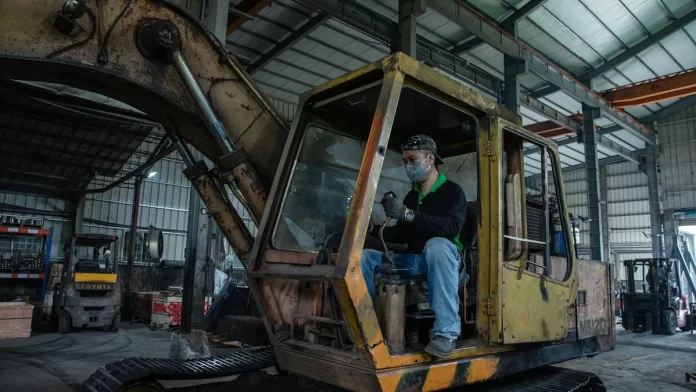 przeglad-koparki-plac-budowy Mechanik w hali serwisowej wykonuje przegląd zużytej koparki gąsienicowej.